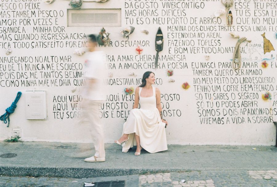 Mulher sentada diante de um muro com poemas escritos, enquanto um vulto passa desfocado